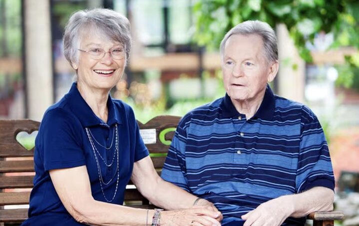 A senior woman and a senior man sitting together on a brown wooden bench outdoors at Mission Grove at Stoneybrook.